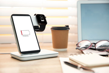 Mobile phone and smartwatch with wireless charger on wooden table. Modern workplace accessory