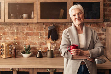 Happy elderly woman standing in kitchen with cup of coffee