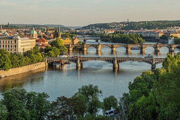 Fototapeta na wymiar Prague / Czech Republic - May 23 2019: Scenic view of the cityscape, the river Vltava and Charles bridge, Manes and Jirasek bridges across water. Green trees in foreground. Sunny spring evening.