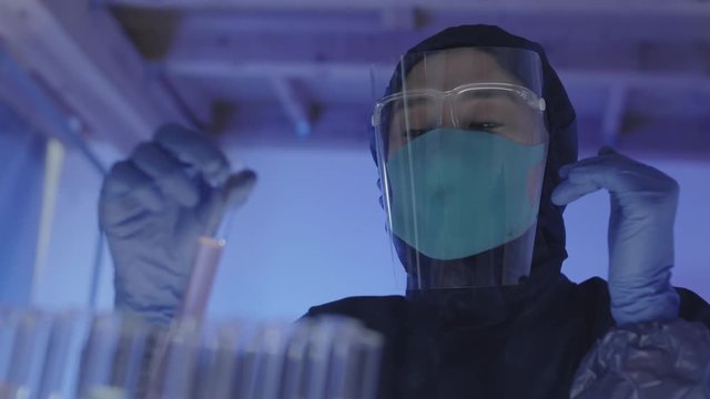 Close up of female Asian microbiologist wearing safety coverall and mask standing in laboratory and examining test-tubes