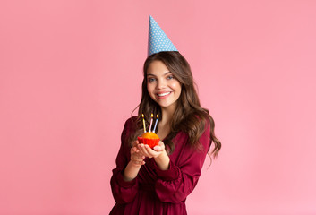 Happy girl in party hat holding birthday cupcake with lighted candles