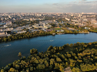 Fototapeta premium A motor boat floats along the coast of the Moscow River, in the background the city outskirts with buildings and roads, beautiful green nature around. Aerial photography