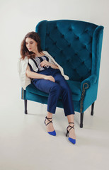 A woman dressed in a fashionable white jacket, striped shirt, blue trousers and shoes on a blue armchair on a white background in the studio.