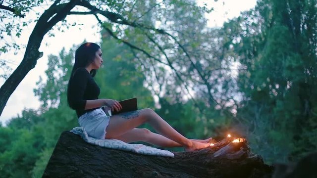 Charming sexy young brunette woman sits on trunk of fallen tree and reads book and near her feet burning candles in forest in evening time
