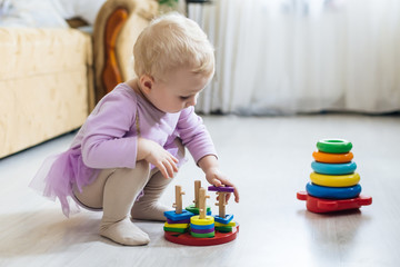 girl plays logical pyramid on floor in living room on sunny day. Montessori wooden toy folded pyramid. Circle, quadra, triangle, rectangle wooden elements of children's toys.