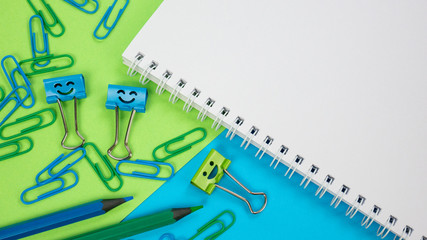 Green and Blue Office Paper Clip, Smile Binder Clips and Pencils on Notepad. Office supplies on Green and Blue Background. Open spiral notebook on table. Knowledge or education. Back to school