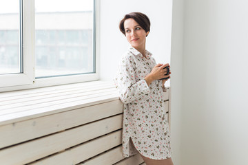 Calm and coziness. Beautiful young woman with cup of tea standing near the window at home
