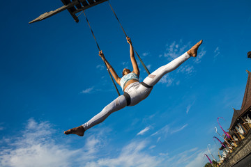 outdoors portrait of young happy and athletic Asian Indonesian woman doing aero yoga workout training body balance  hanging from swing rope isolated on blue sky