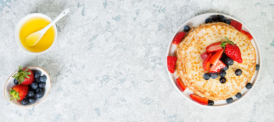 A stack of crepes lies in a plate, decorated with berries on a gray concrete background. Top view, copy of space.