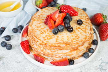 A stack of crepes lies in a plate, decorated with berries on a gray concrete background