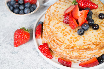 A stack of crepes lies in a plate, decorated with berries on a gray concrete background.