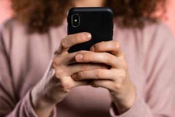 Hands of girl with curly hair using smartphone or surfing internet on pink studio background. Modern technology - apps, social networks