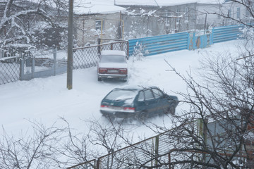 Rural snowy street. Car driving along a a winter road. Another car is parked near the courtyard of the house.