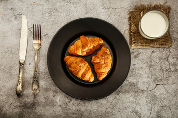 Freshly baked French croissants on a black plate with a fork and knife on a gray concrete background. View from above.