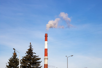 Industrial chimney with smoke and treetops against blue sky