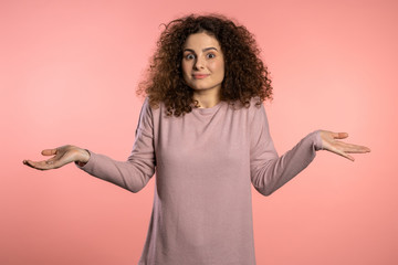  Young curly unsure girl shrugs her arms, makes gesture of I don't know, care, can't help anything .Young curly woman with trendy glitter freckles make-up