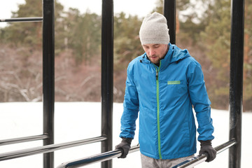 Concentrated young man in hat and jacket looking down while keeping body on parallel bars