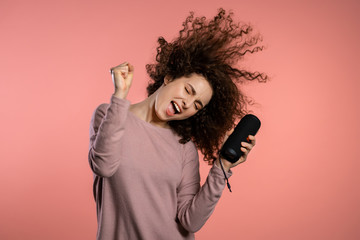 Cute woman with curly hair smiling, dancing with wireless portable speaker in studio on pink background. Music, dance concept