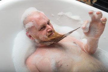 A bald man is fooling around in the foam bath. The guy makes funny hairstyles on his red beard.