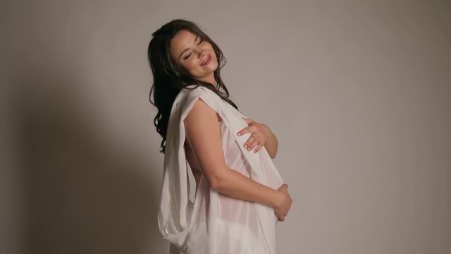 Beautiful naked pregnant woman with white silk on white background, pregnancy nude concept. Pregnant woman touching her belly with hands.