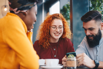 Three friends sitting in a cafe, talking, drinking coffee and looking at the male friend's phone.