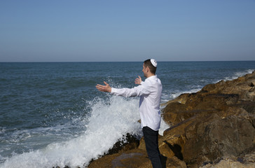 young Jew stands with arms outstretched in meditation on a rocky seashore with a large wave hitting the rocks at his feet.