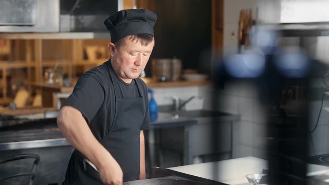 Busy Asian kitchener worker in black clothing cooking food in frying pan. Medium shot on RED camera