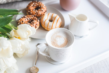 Breakfast in bed, cup with cappuccino, doughnuts, flowers white tulips, morning concept, linen bed sheets, woman's day