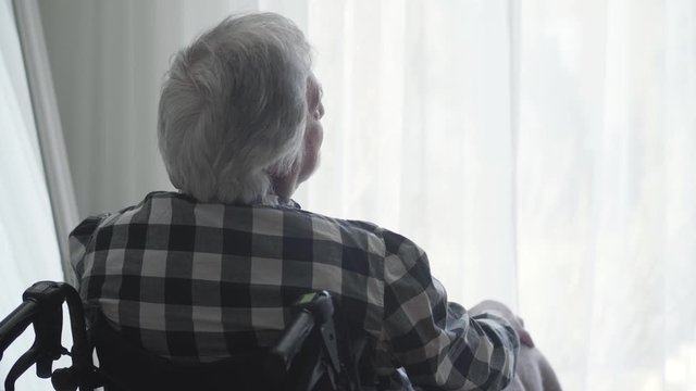 Close-up back view of lonely elderly Caucasian man sitting in wheelchair in front of big window closed with curtain and thinking. Lonely mature man spending day alone at home. Oldness, disability.