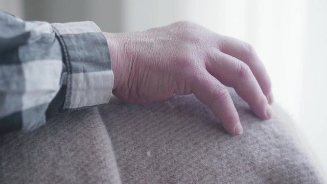 Extreme close-up of old male Caucasian hand on knees covered with blanket. Elderly ill man sitting alone indoors. Health care, retirement, disability.