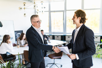 Businessmen shaking hands. Two confident businessmen shaking hands and smiling while standing at the meeting together with people in the background