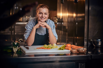European female chef standing in a dark kitchen next to cutting board with vegetables on it,...