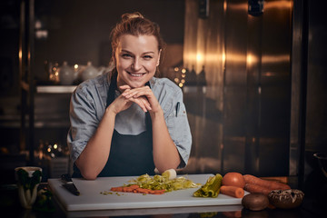 European beautiful female chef standing in a dark kitchen next to cutting board with vegetables on...