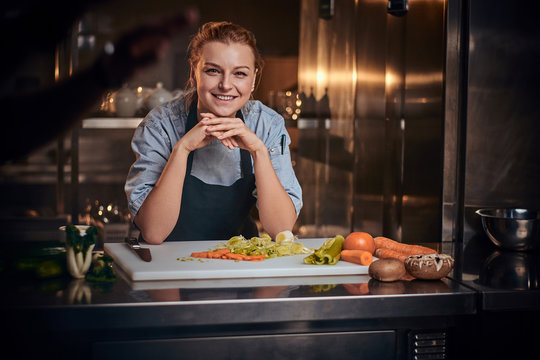 European female chef standing in a dark kitchen next to cutting board with vegetables on it, wearing apron and denim shirt, posing for the camera, reality show look