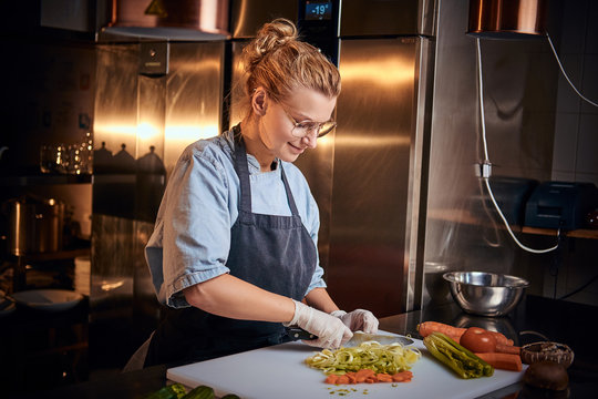 Smiling And Calm Woman Chef Standing In A Dark Restaurant Kitchen Next To Cutting Board While Cutting Vegetables On It, Wearing Apron And Denim Shirt, Posing For The Camera, Reality Show Look