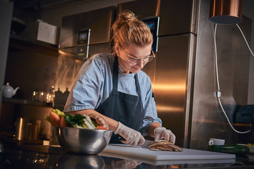 Interested and serious female chef standing in a dark restaurant kitchen next to cutting board while cutting vegetables on it, wearing apron and denim shirt, posing for the camera, cooking show look