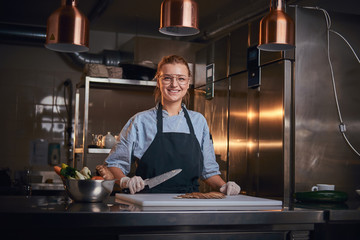 Joyful and excited female chef standing in a dark cafe kitchen next to big bowl of fresh vegetables and cutting board with mushroom pieces on it, holding a knife, wearing apron and denim shirt, posing