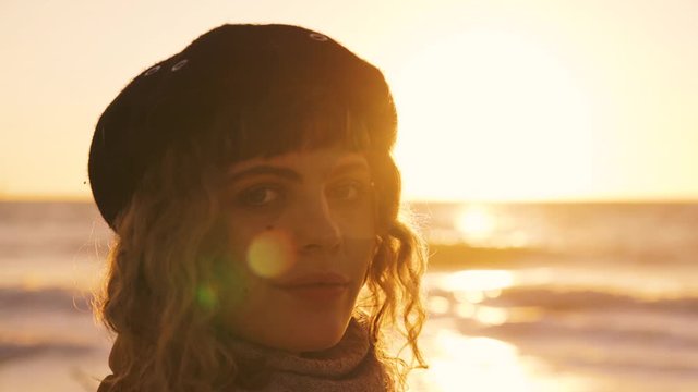 Slow-motion of a young brunette model girl with a fringe and a black vintage basque cap at a beach in Florida by the golden light of the evening sun with flares by sunset making eye contact and waves