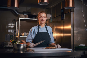 Confident and serious woman chef standing in a dark kitchen next to a big bowl of fresh vegetables and cutting board with mushroom pieces on it, holding a kitchen knife, wearing apron and denim shirt