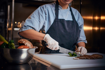 Chef female standing on a dark restaurant kitchen, wearing gloves, apron and denim shirt, cutting vegetables on a cutting board net to the bowl with raw vegetables, close up view