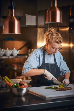 Calm and serious female chef standing in a dark restaurant kitchen next to big bowl of vegetables and a cutting board while slicing pickles, wearing apron, denim shirt, and glasses, posing for the
