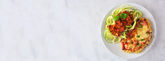 Cauliflower parmesan and zucchini pasta topped with meatless walnut cauliflower bolognese. Top view on a white marble banner background. Healthy eating, plant-based meat substitute concept.
