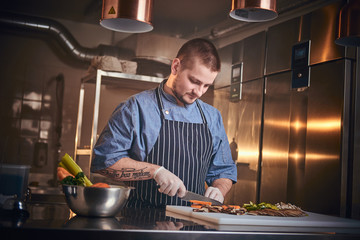 Confident and serious male chef standing in a dark kitchen next to cutting board with vegetables on...