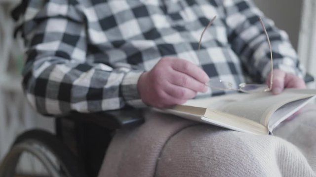 Unrecognizable old caucasian man holding book and eyeglasses as sitting in wheelchair. Disabled retiree reading at home. Hobby, retirement, disability, lifestyle.