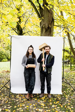 Portrait of woman and bearded man holding wooden blocks standing in front of white background in a garden, looking at camera.