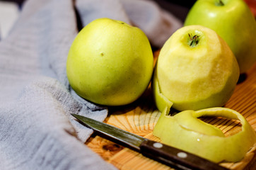 Three fresh green apples lie on a wooden cutting Board, along with a gray linen napkin and a knife. One of the apples has been peeled off. Selective focus. Close up.