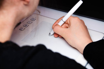 Female hands work on a graphic tablet. Hand holds stylus pen and draws. White graphic tablet. The work of a graphic designer. Girl works on a tablet connected to a laptop. Rear view from  behind