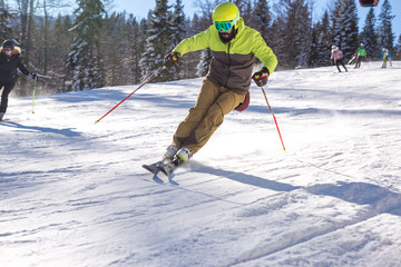 On a sunny morning on the mountain, a Skier in a yellow jacket goes down the slope. The cable car is just above the ski slope and pine trees are all around.