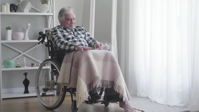 Long shot, nervous old Caucasian disabled man beating fist on painful knee. Mature ill retiree sitting in wheelchair indoors. Disease, disability, injury, paralysis.