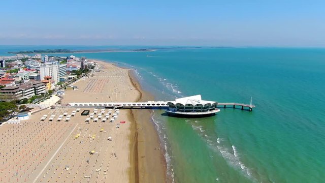 Lignano, Italy famous beach pier. Sunny summer day. Golden sand beach. Parallax aerial shot.